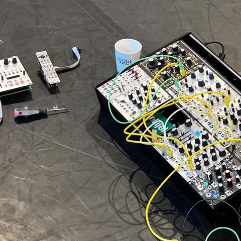 a photo of my synthesizer with a few modules removed, placed on a linoleum dance floor. In the frame, there’s also a club guy and a Roni coffee cup. It’s one of the first pictures I took while rehearsing and configuring the system I used to create music with the dancers for the project.